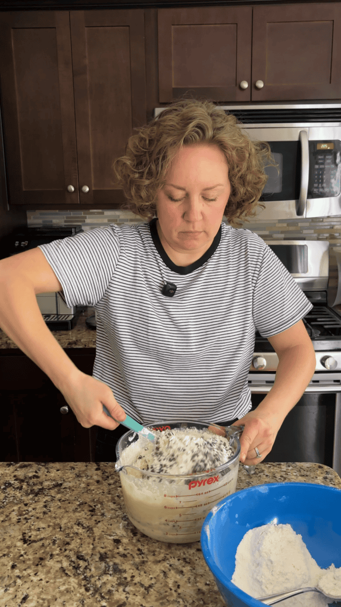 A person with curly hair in a striped shirt mixes ingredients in a large glass bowl on a granite kitchen counter, preparing coconut chocolate chip muffins. There is flour in a blue bowl nearby and kitchen appliances in the background.