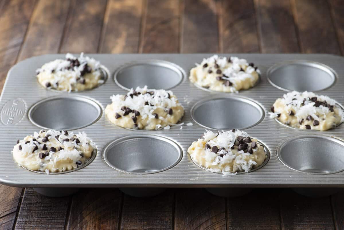 A muffin tin with five unbaked coconut chocolate chip muffins, each generously topped with shredded coconut and chocolate chips, sits on a wooden surface; four spaces in the tin are empty.
