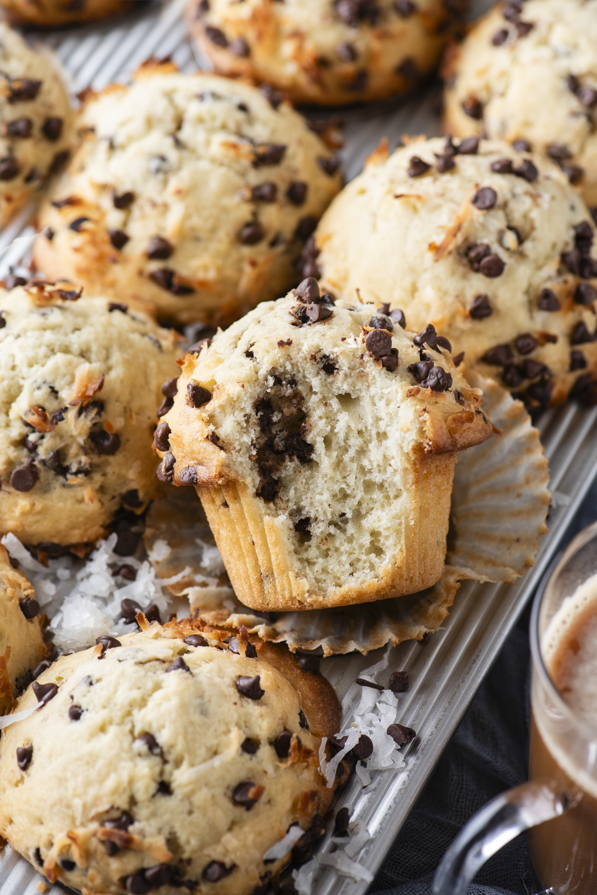 A tray of coconut chocolate chip muffins, one unwrapped with a bite taken out, reveals a soft and tender interior. Shredded coconut and mini chocolate chips are sprinkled on top, with a glass of hot cocoa nearby.