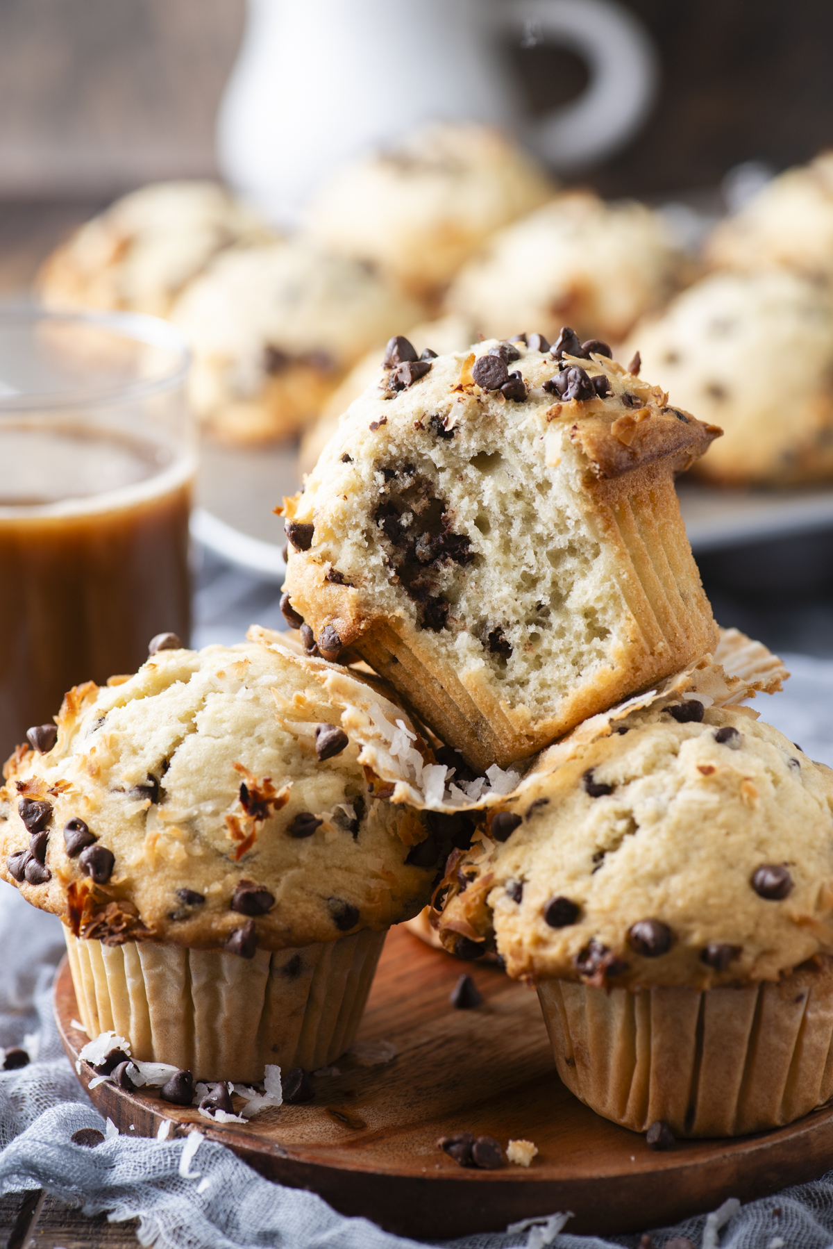 Three coconut chocolate chip muffins on a wooden board, one split in half to show the soft, coconut-studded inside, with a glass of coffee and more muffins blurred in the background.