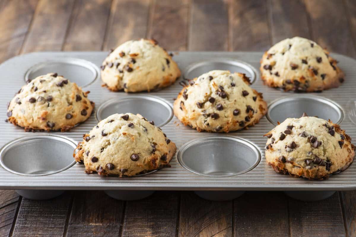 A muffin tin with seven freshly baked coconut chocolate chip muffins, each golden brown and studded with chocolate chips, sits on a wooden surface.
