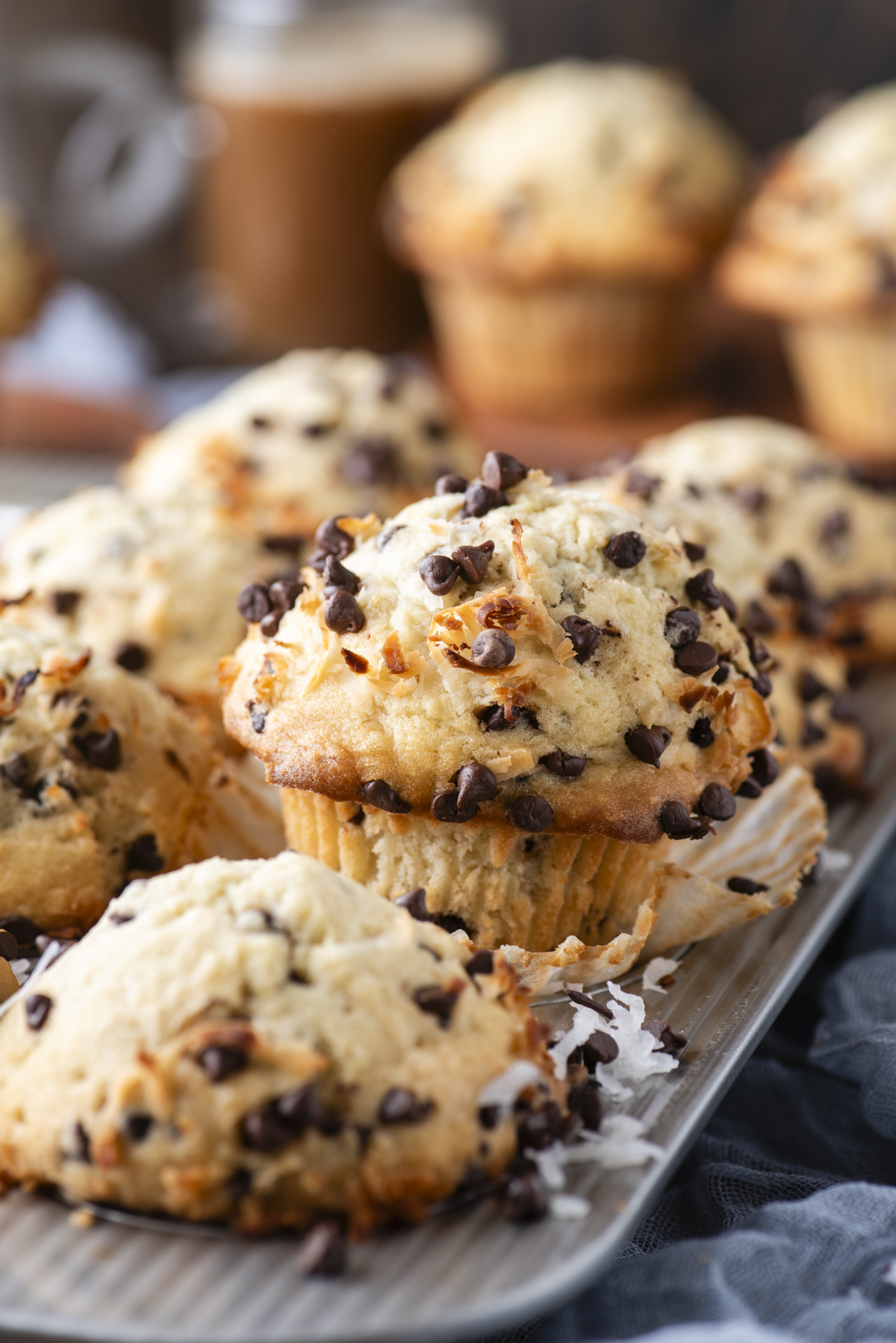 A tray of freshly baked coconut chocolate chip muffins with chocolate chips on top, with one muffin unwrapped in the center. More muffins and a blurred drink are visible in the background.