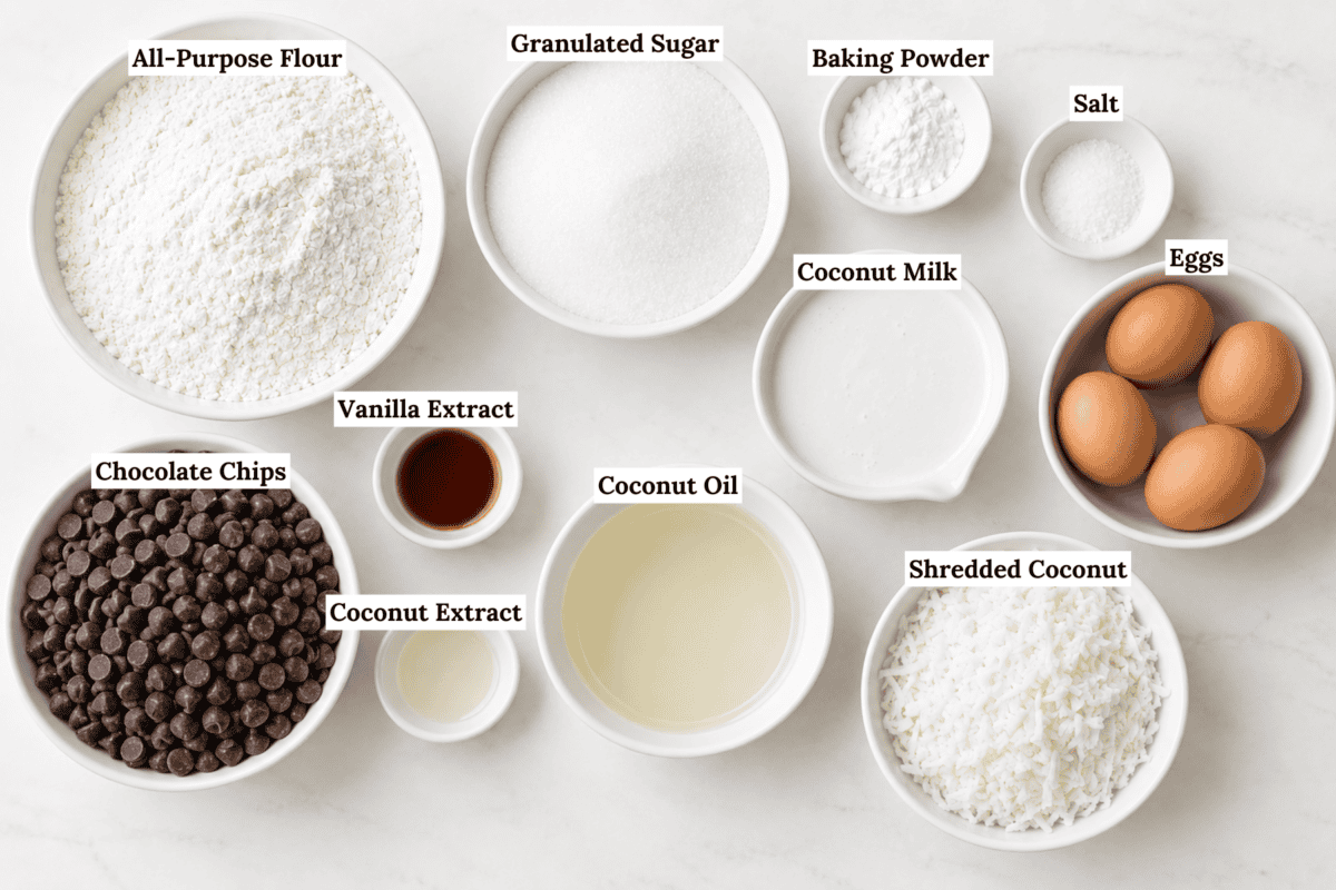 Multiple labeled bowls on a white surface contain ingredients perfect for coconut chocolate chip muffins: all-purpose flour, granulated sugar, baking powder, salt, eggs, coconut milk, coconut oil, shredded coconut, chocolate chips, vanilla extract, and coconut extract.