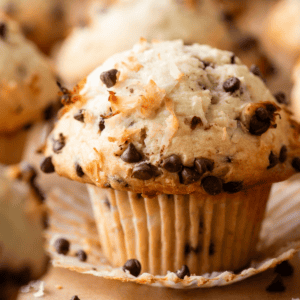 A close-up of one of our coconut chocolate chip muffins with a crinkled paper liner, topped with shredded coconut and chocolate chips, surrounded by more muffins in soft focus.