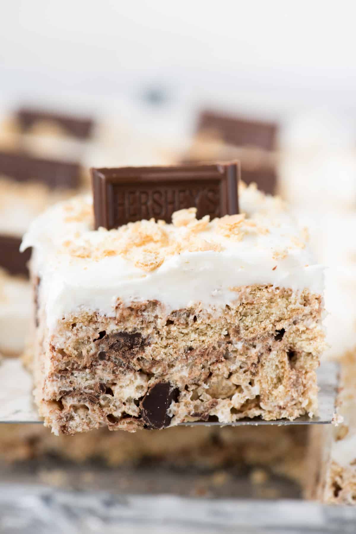Rice krispie s’mores treat with marshmallow frosting topped with hershey’s bar on metal serving utensil 