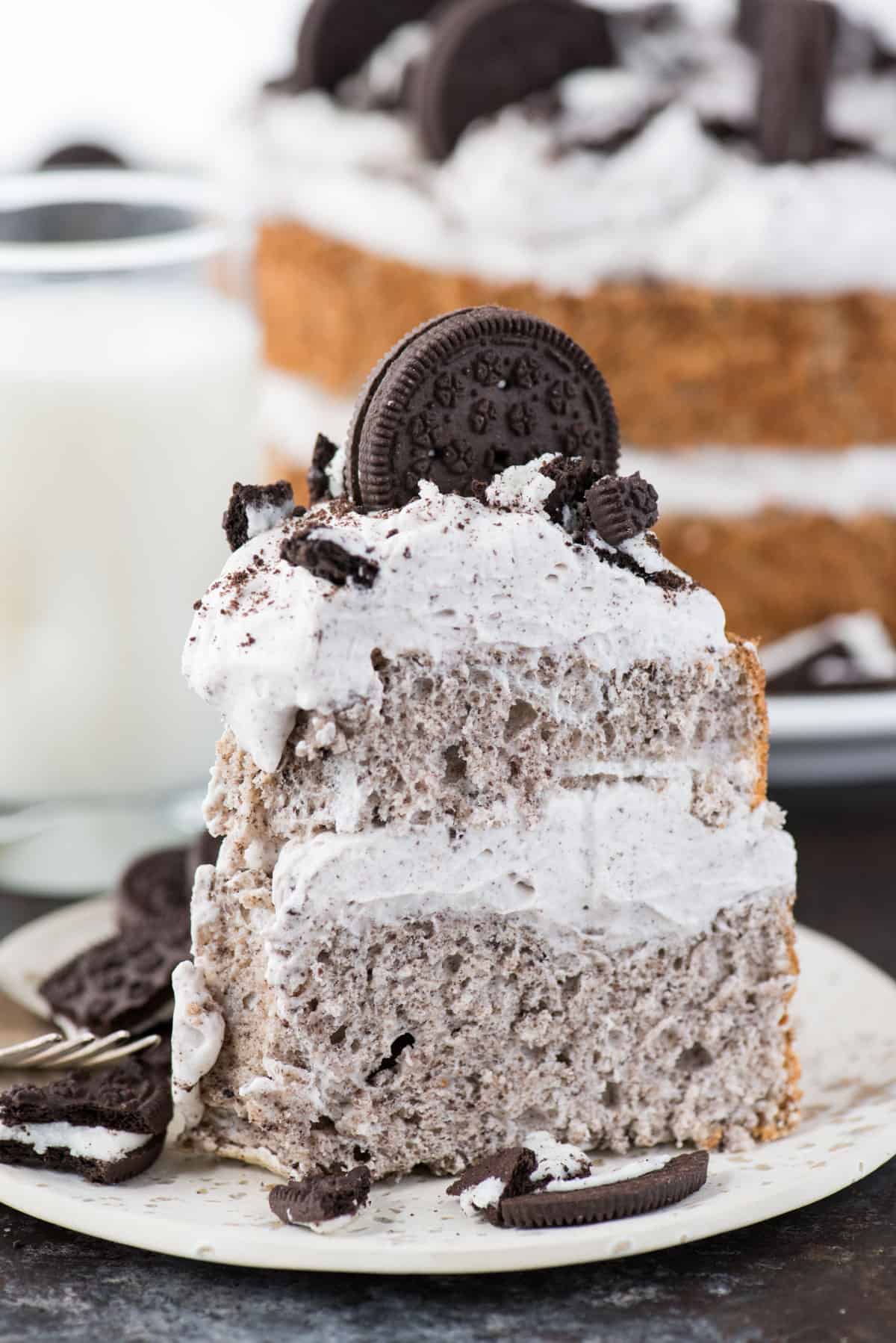 slice of oreo angel food cake with oreo whipped cream and crushed oreo pieces on white plate on dark background