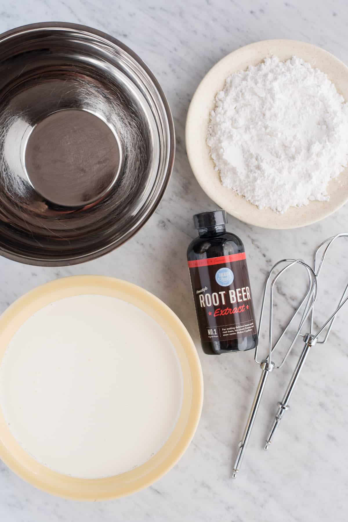 ingredients to make root beer frosting, includes metal bowl, powdered sugar, heavy cream, root beer extract and mixing beaters