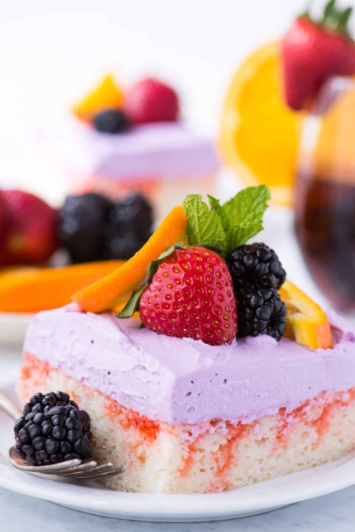 slice of sangria poke cake garnished with a strawberry, blackberries, orange slice and mint leaf on white plate