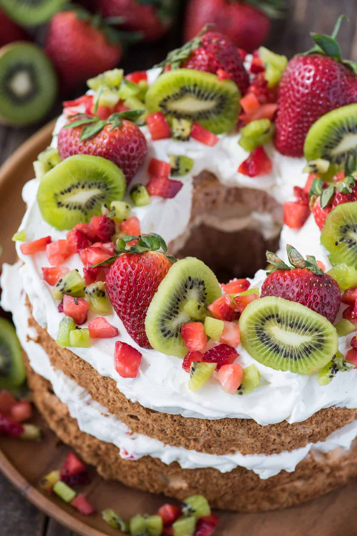 strawberry angel food cake topped with whipped cream, whole strawberries, chopped strawberries and kiwi on wooden serving tray
