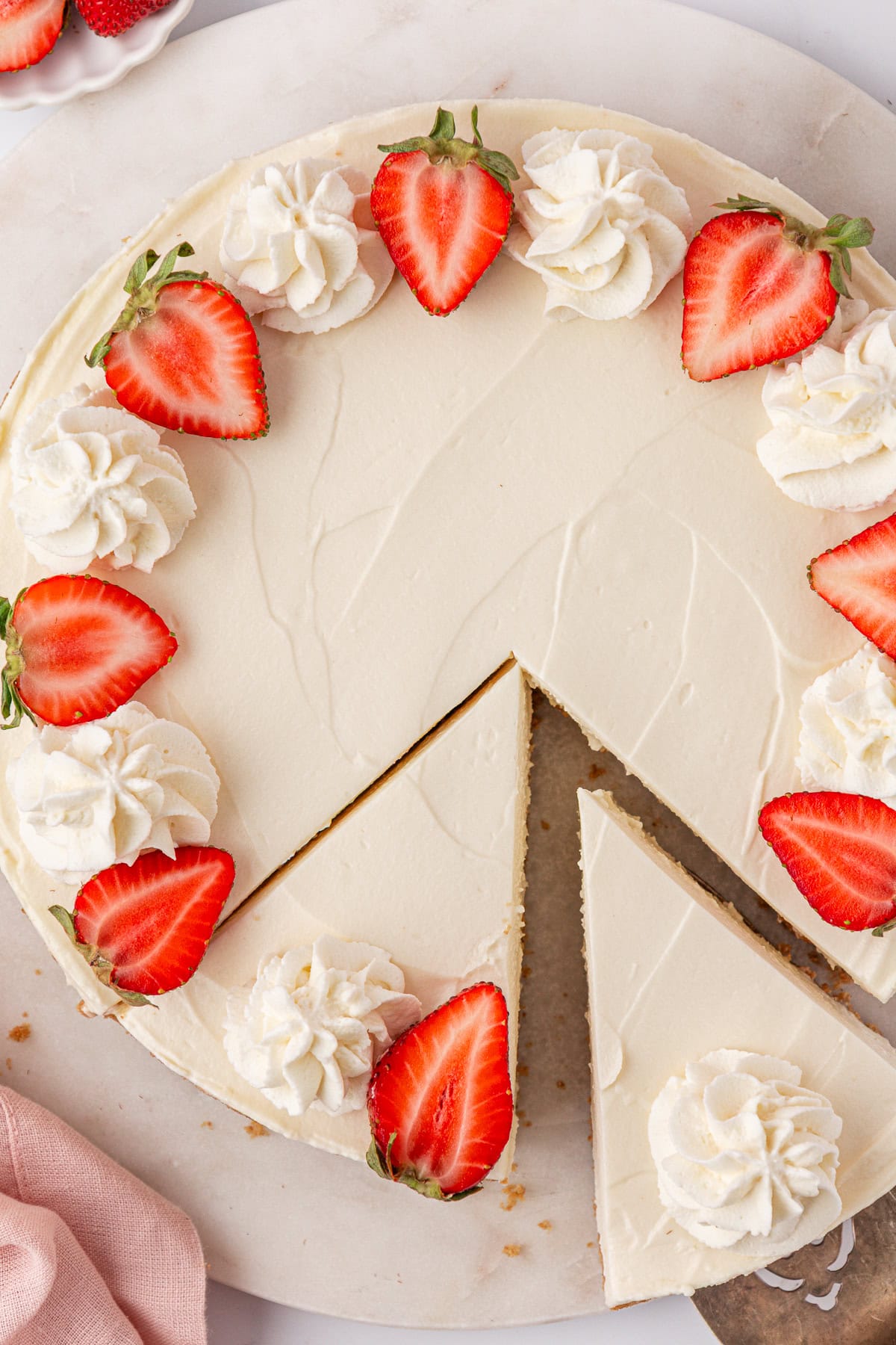 A round Philadelphia no bake cheesecake topped with whipped cream swirls and halved strawberries, with one slice being lifted out. The cake sits on a white surface next to a pink cloth.