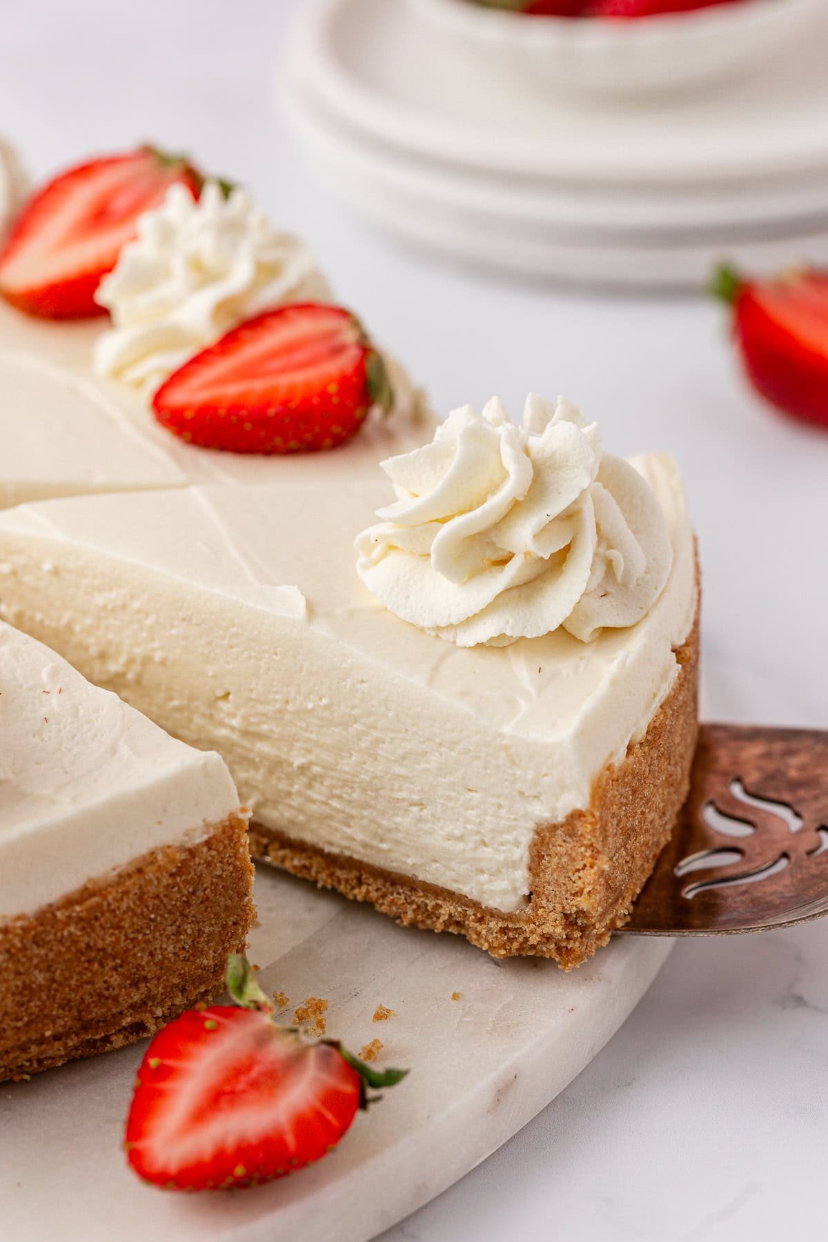 A slice of creamy Philadelphia no bake cheesecake with a graham cracker crust is being served. Topped with swirls of whipped cream and halved strawberries, this delightful dessert sits on a pristine white surface.