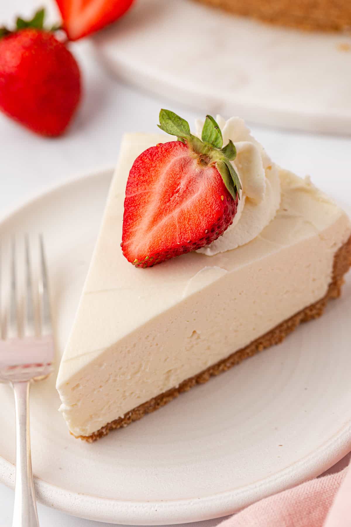 A slice of creamy Philadelphia no bake cheesecake with a graham cracker crust, topped with whipped cream and half a fresh strawberry, served on a white plate with a fork beside it.