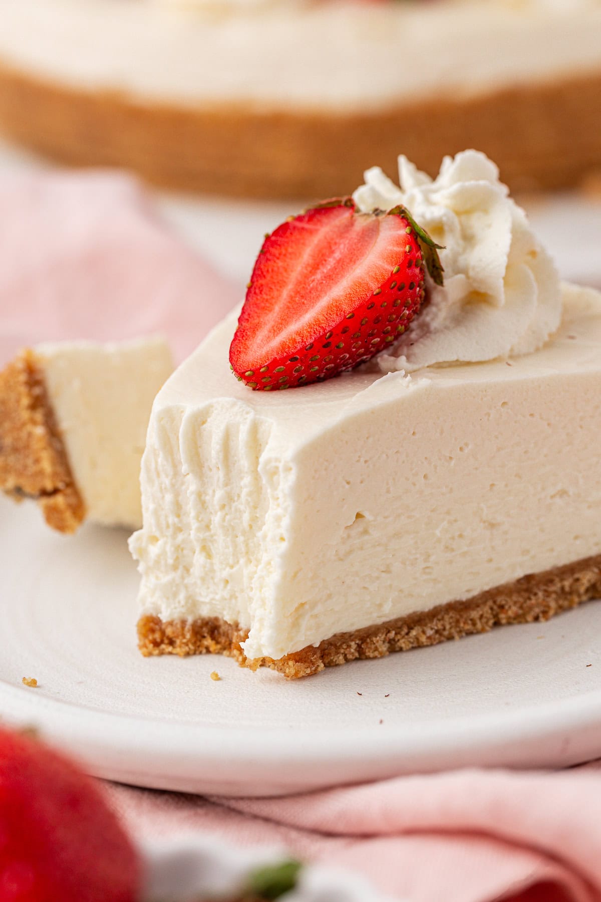 A creamy slice of Philadelphia no bake cheesecake with a graham cracker crust, topped with a halved strawberry and a swirl of whipped cream, sits on a white plate with a blurred background.