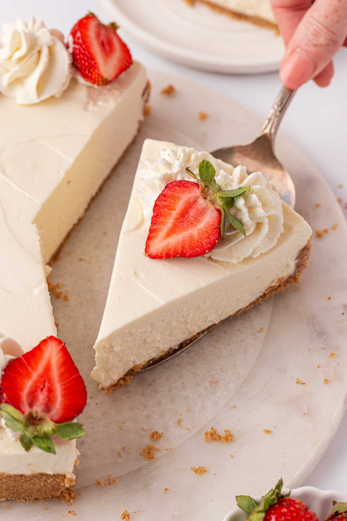 A hand lifts a slice of creamy Philadelphia no bake cheesecake topped with whipped cream and a halved strawberry from a whole cheesecake on a marble surface. Crumbs and more strawberries are visible nearby.