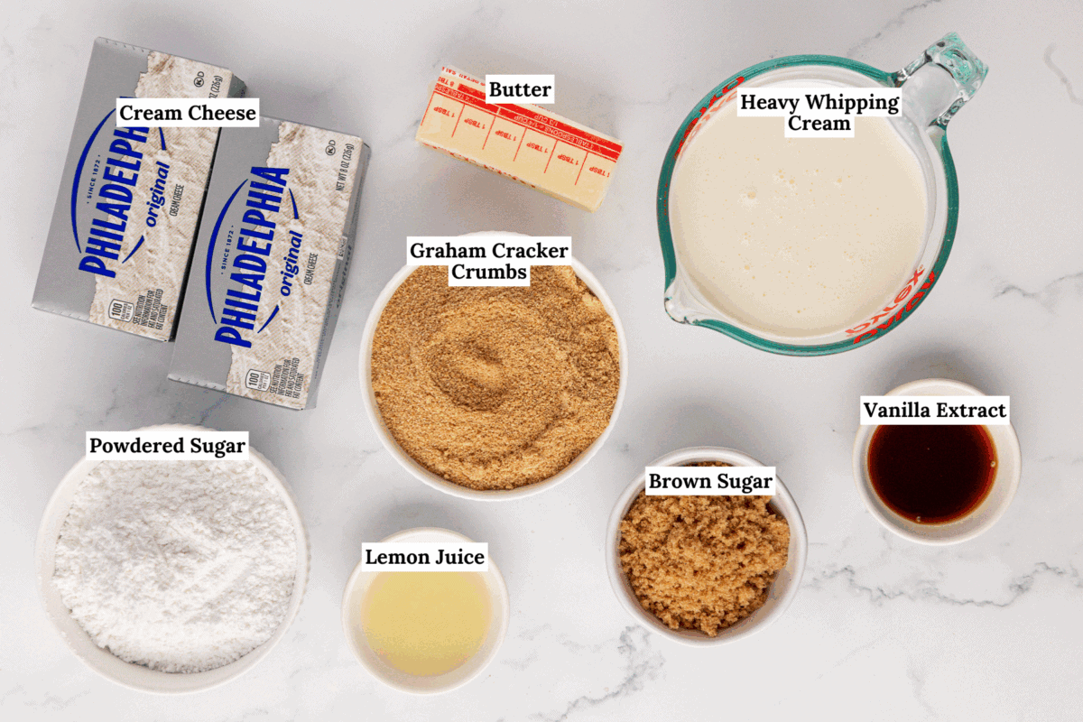 Ingredients for a Philadelphia no bake cheesecake are arranged on a white surface: cream cheese, butter, heavy whipping cream, graham cracker crumbs, powdered sugar, lemon juice, brown sugar, and vanilla extract, each labeled.