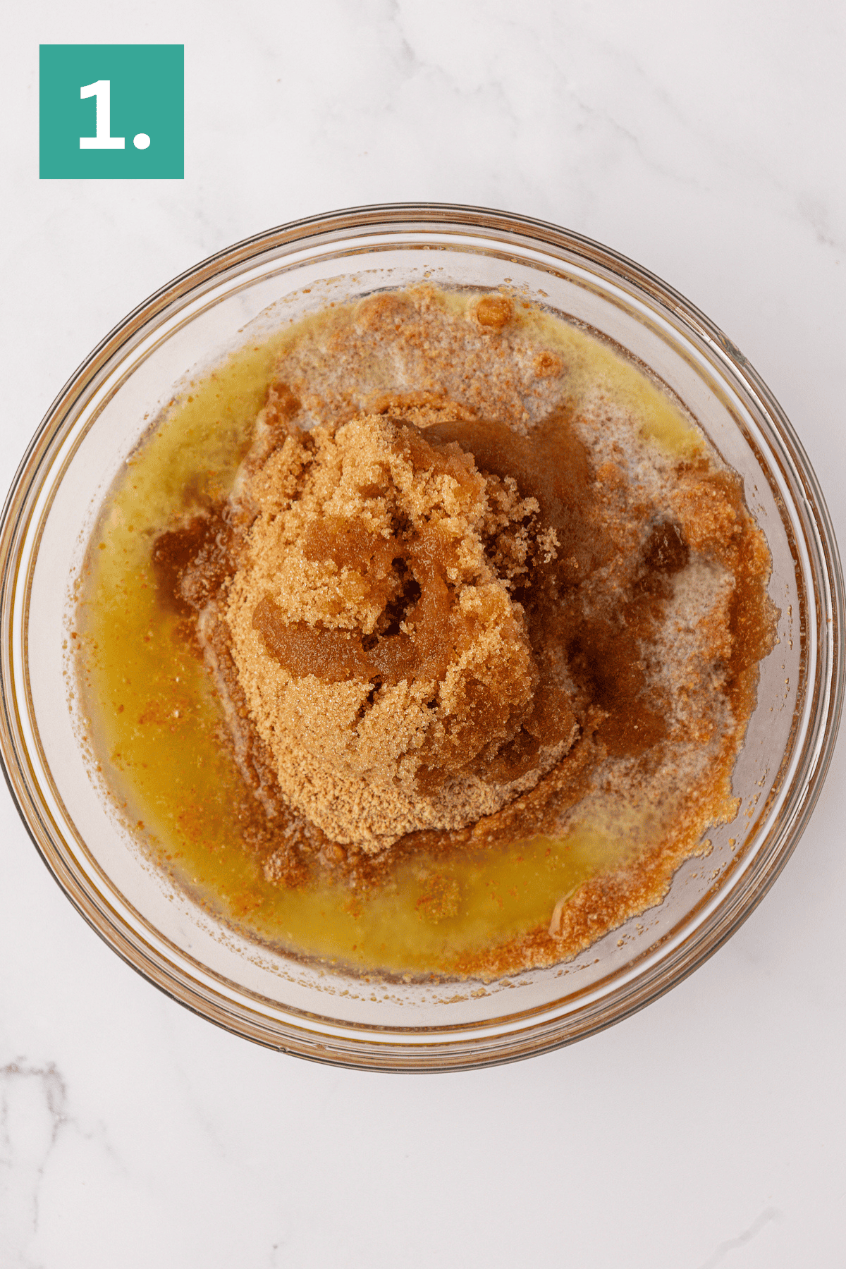 A glass bowl filled with melted butter, brown sugar, and cinnamon sits on a white marble surface. A green box with the number 1 appears in the top left corner, marking the first step for making a Philadelphia no bake cheesecake.