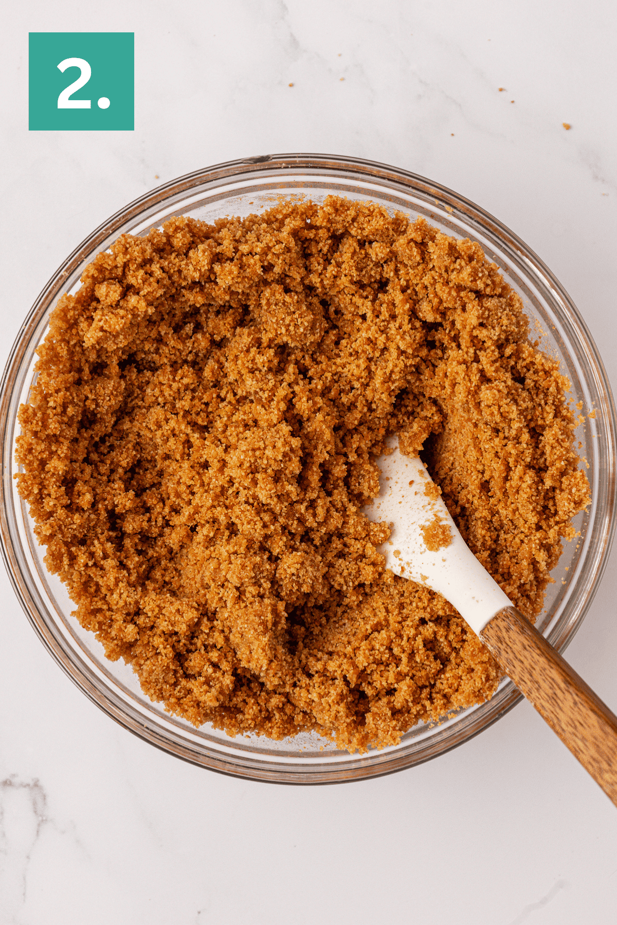 A glass bowl filled with a crumbly brown mixture for a Philadelphia no bake cheesecake sits beside a white spatula with a wooden handle. A green square with the number 2 appears in the top left corner.