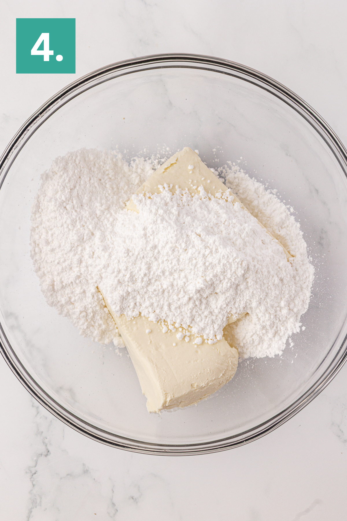 A glass mixing bowl with cream cheese and powdered sugar, perfect for making Philadelphia no bake cheesecake, sits on a white surface. A green square with the number 4 in white is in the top left corner.