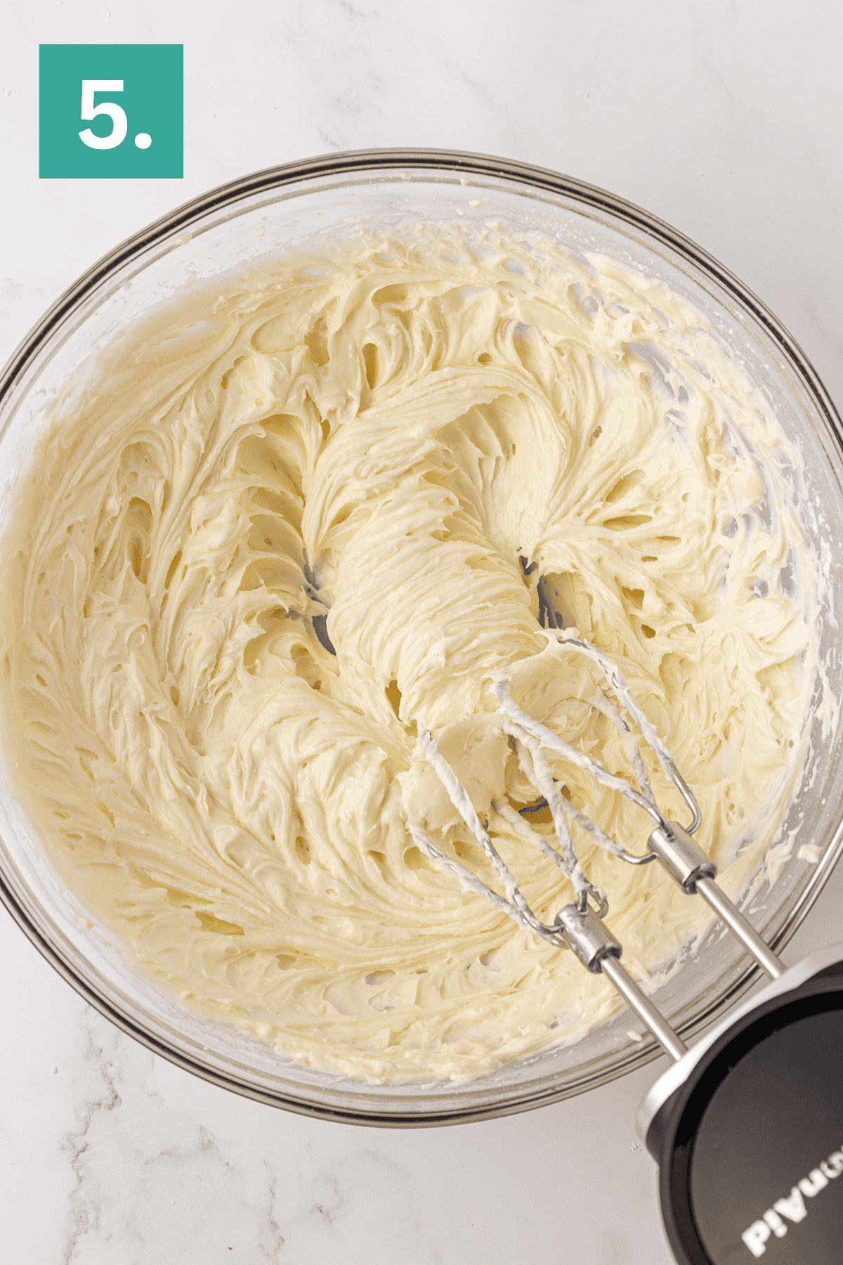 A glass bowl of whipped cream cheese mixture for a Philadelphia no bake cheesecake is being mixed with an electric hand mixer, with a green box labeled 5. in the top left corner.