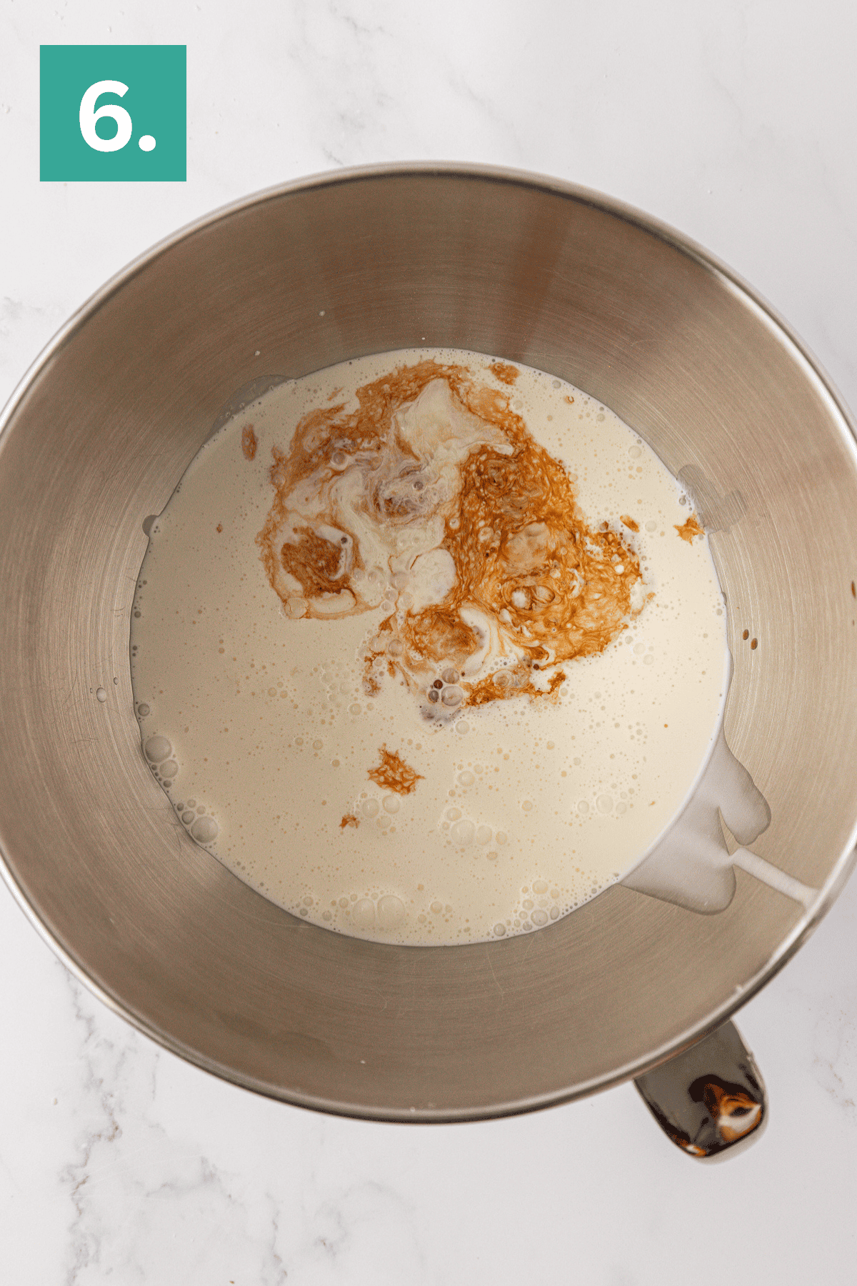 A metal mixing bowl containing cream and vanilla extract for a Philadelphia no bake cheesecake, shown from above on a white marble surface. A green box with the number 6 is in the top left corner of the image.