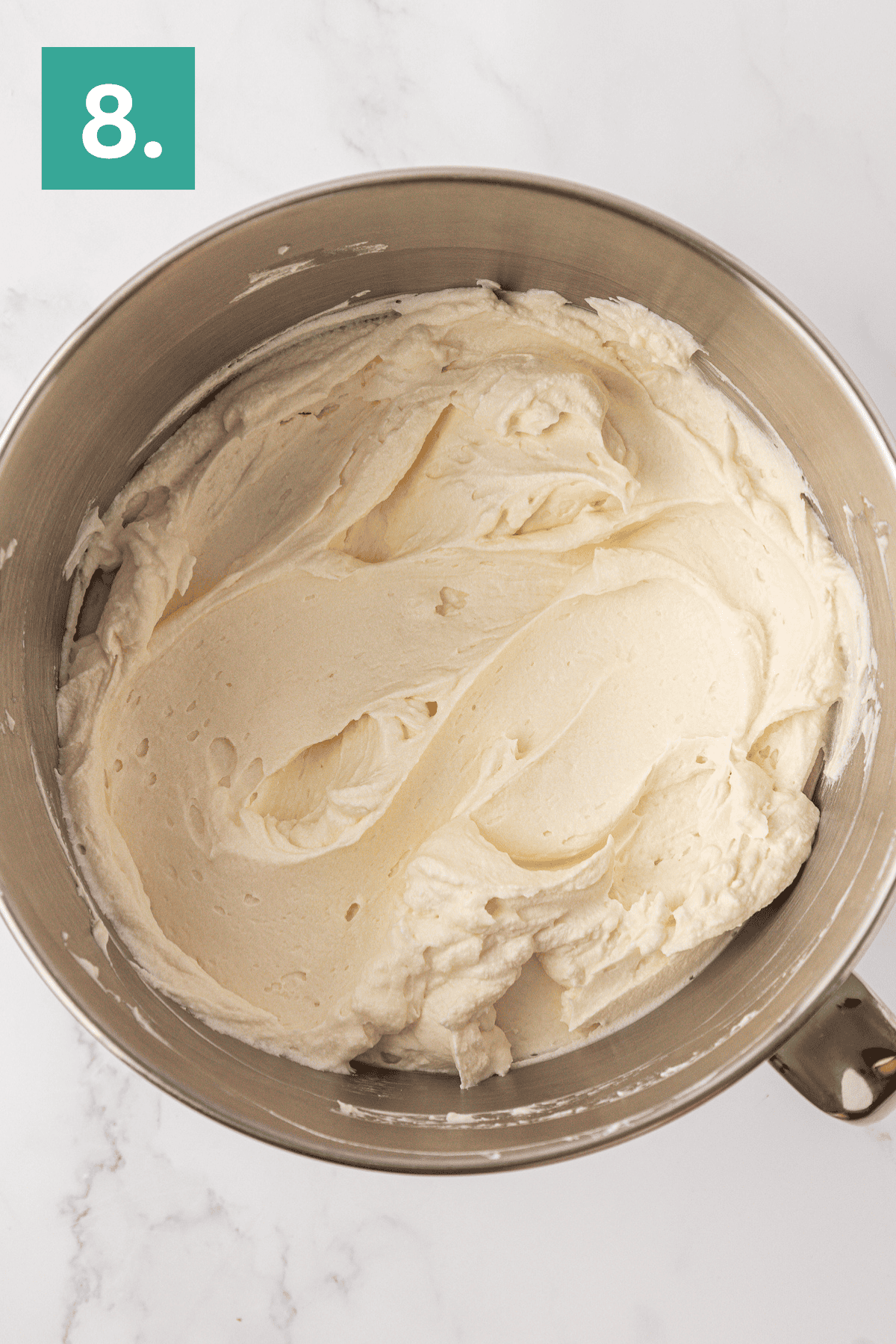A mixing bowl filled with smooth, creamy white frosting for a Philadelphia no bake cheesecake. In the top left corner, a green box with the number 8 is displayed. The background is a white marble surface.