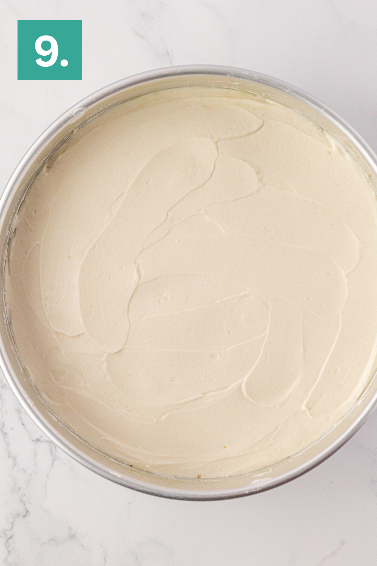 A round cake pan filled with a smooth, evenly spread Philadelphia no bake cheesecake batter sits on a marble surface. A green square with the number 9 is in the top left corner.