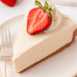 A slice of creamy Philadelphia No Bake Cheesecake with a crumbly crust, topped with whipped cream and a halved strawberry, served on a white plate with a fork beside it.