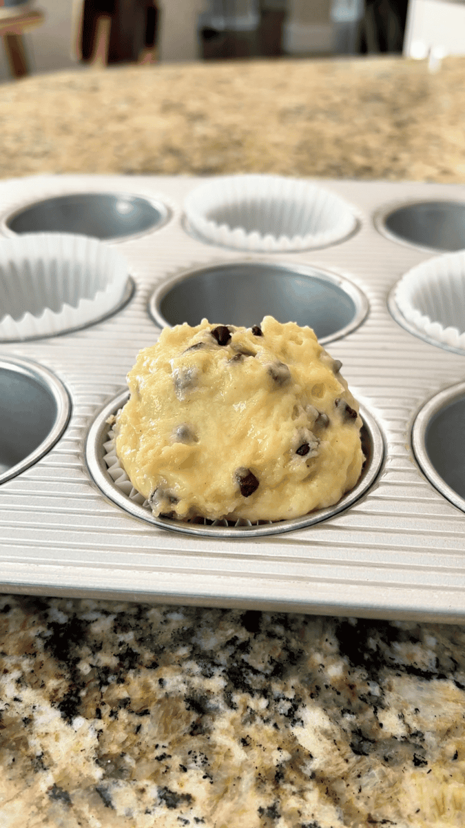 A muffin tin lined with paper cups holds one scoop of chocolate chip muffins batter in the foreground, resting on a granite countertop.