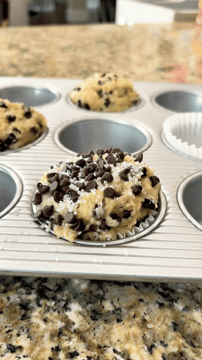 A muffin pan filled with raw chocolate chip muffin batter in paper liners, these irresistible chocolate chip muffins are topped with mini chocolate chips and a sprinkle of sugar, ready to be baked.