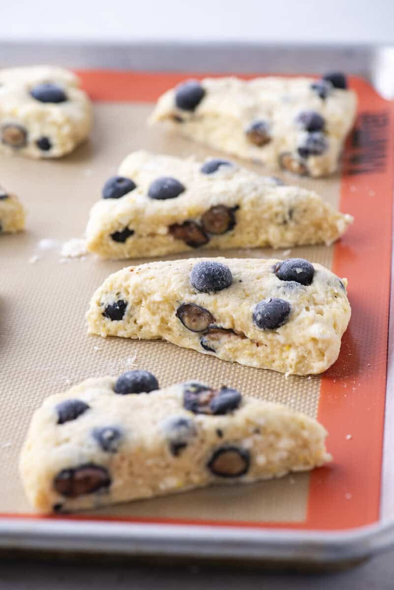 Unbaked lemon blueberry scones are arranged on a baking sheet lined with a silicone mat, ready to go into the oven. The dough is studded with fresh blueberries and lightly dusted with flour.