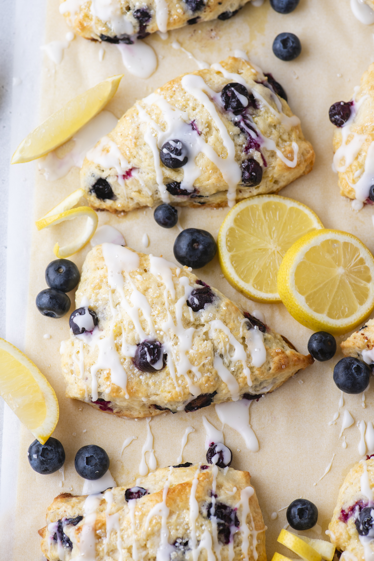 Lemon blueberry scones drizzled with white icing are arranged on parchment paper, surrounded by fresh blueberries and lemon slices.