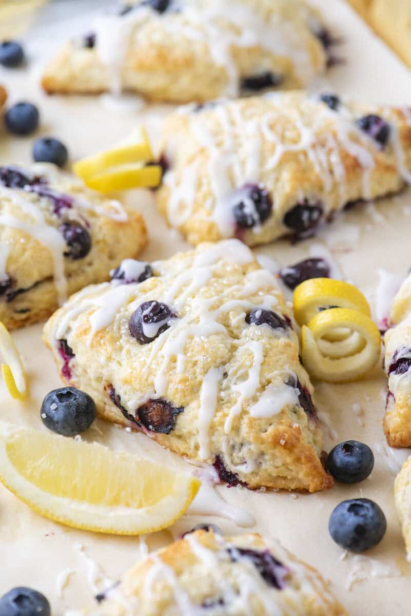 Triangular lemon blueberry scones drizzled with white icing are arranged on parchment paper, surrounded by fresh blueberries, lemon twists, and a lemon wedge.
