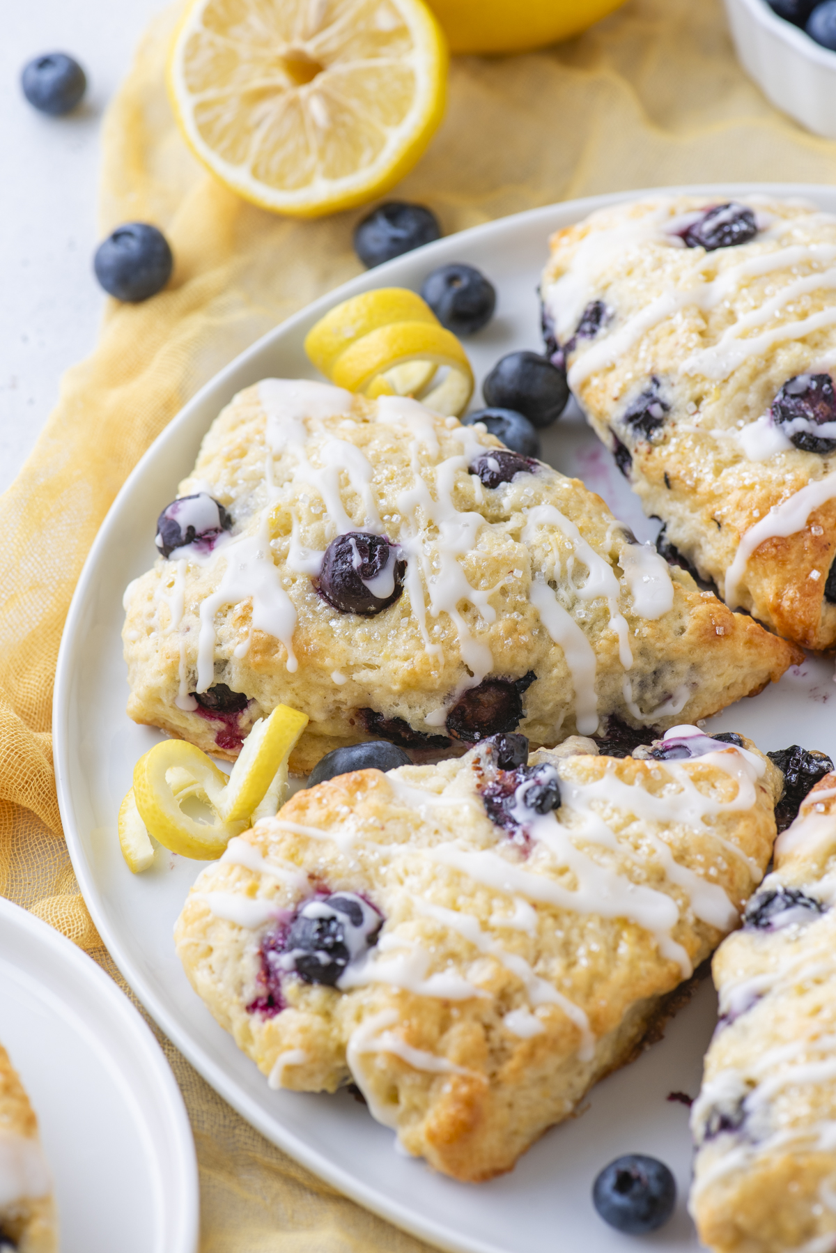 A plate of lemon blueberry scones drizzled with white glaze, garnished with fresh blueberries and lemon twists, sits on a yellow cloth with a sliced lemon in the background.