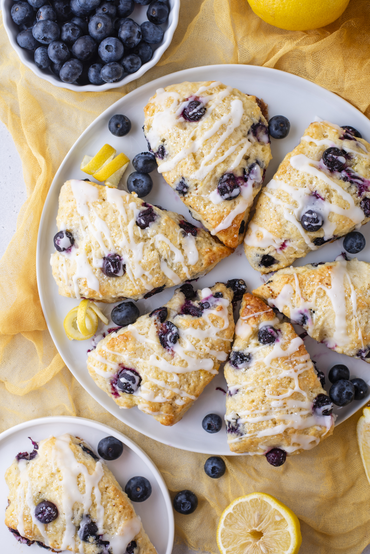 A plate of lemon blueberry scones drizzled with icing, surrounded by fresh blueberries and lemon slices on a yellow cloth, with a bowl of blueberries and a halved lemon nearby.