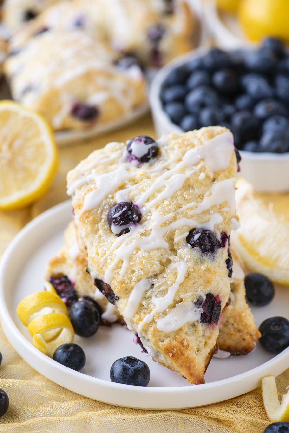 Two triangular lemon blueberry scones drizzled with white icing sit on a white plate, surrounded by fresh blueberries and lemon slices, with more scones and blueberries in the background.
