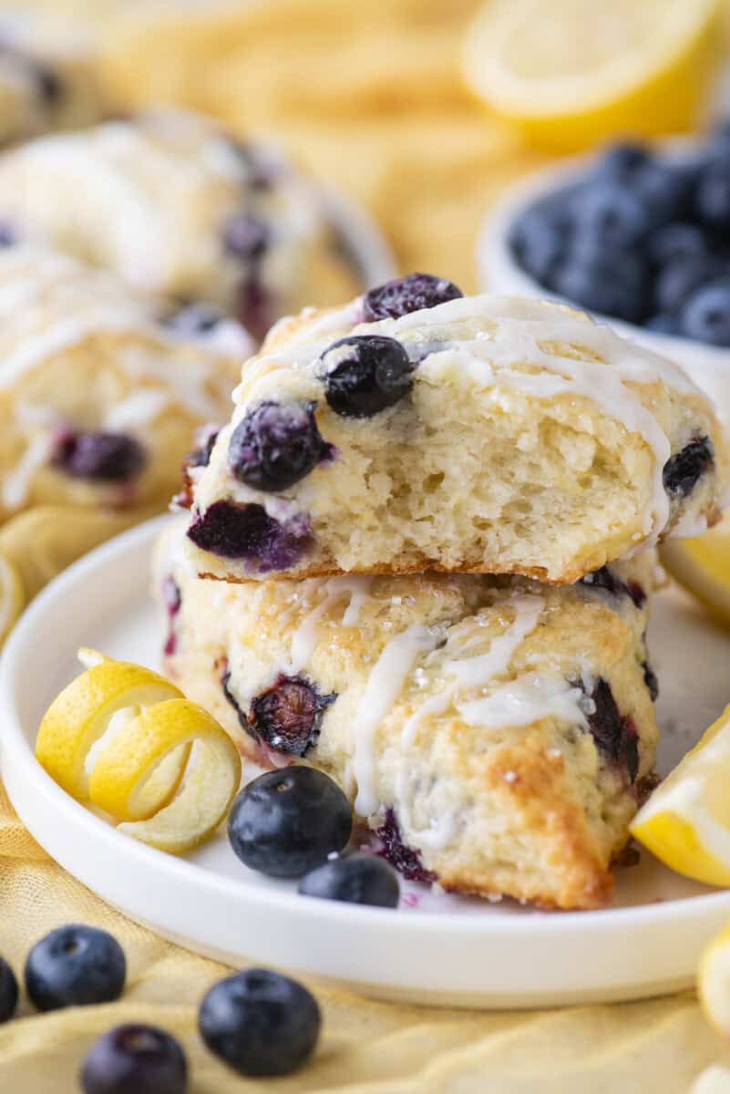 A close-up of two lemon blueberry scones drizzled with white icing on a white plate, surrounded by fresh blueberries and lemon zest curls, with more scones and blueberries in the background.