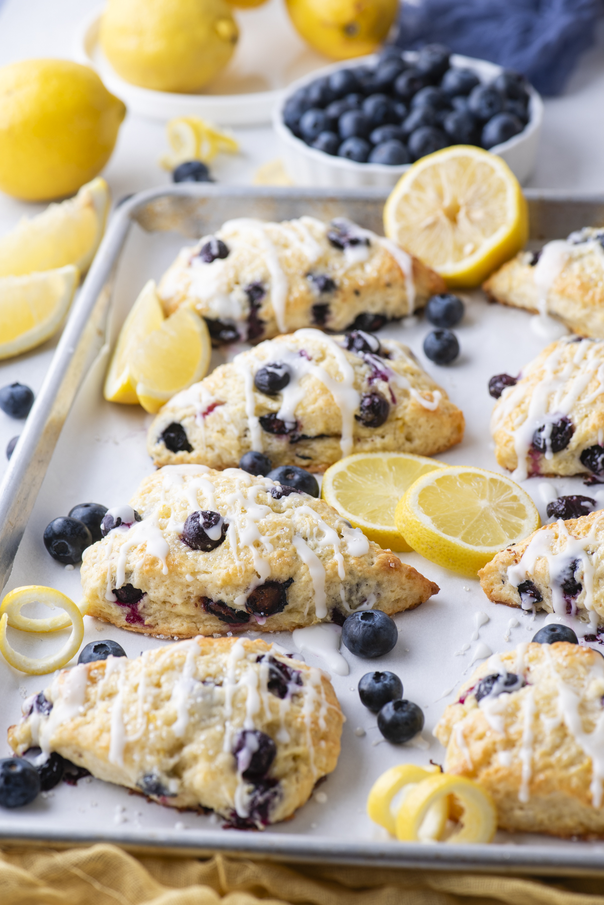 Lemon blueberry scones drizzled with icing are arranged on a baking tray, garnished with fresh blueberries and lemon slices. Lemons and a bowl of blueberries are in the background.