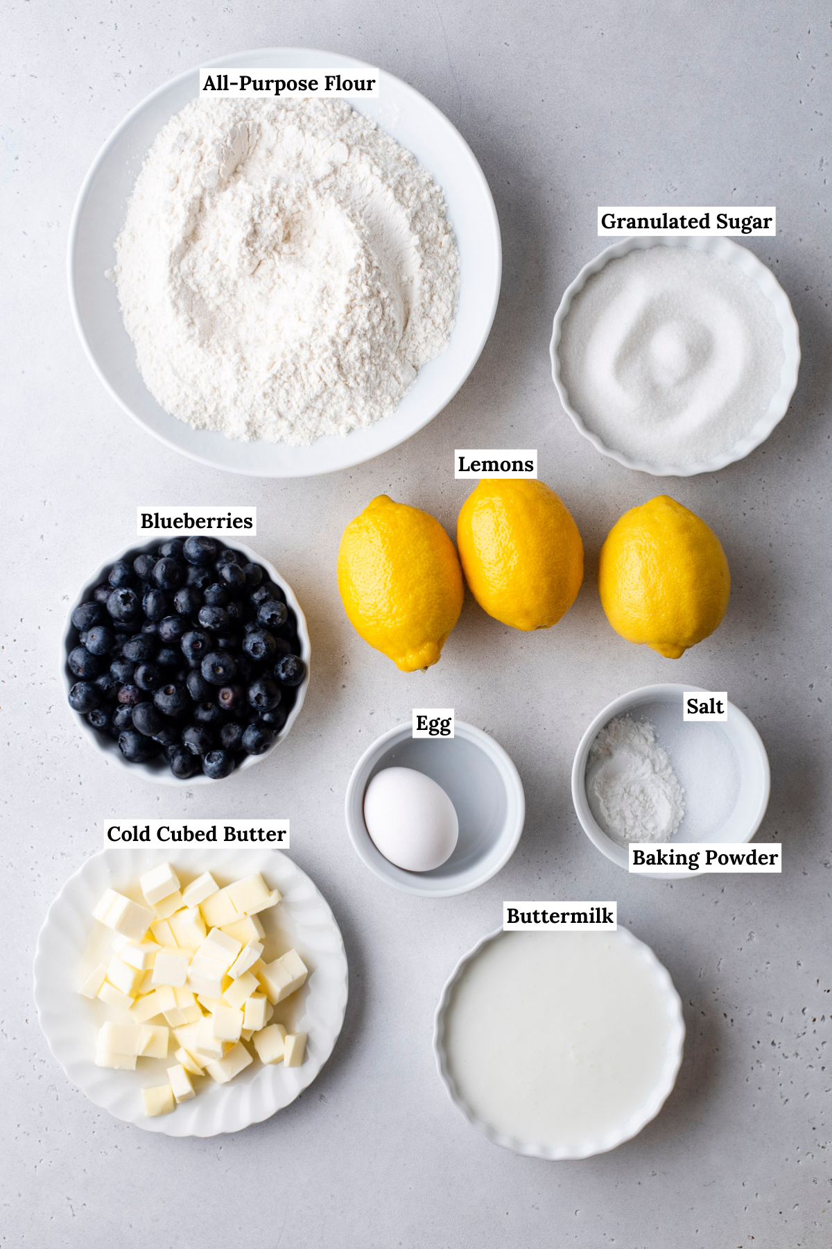 Top-down view of ingredients for lemon blueberry scones: a bowl of flour, sugar, three lemons, blueberries, cubed butter, an egg, salt, baking powder, and buttermilk. Perfect for baking deliciously fresh scones.