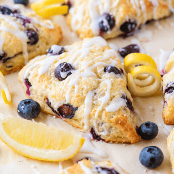 A close-up of a lemon blueberry scone drizzled with white icing, surrounded by fresh blueberries, lemon slices, and lemon zest curls on a light surface.