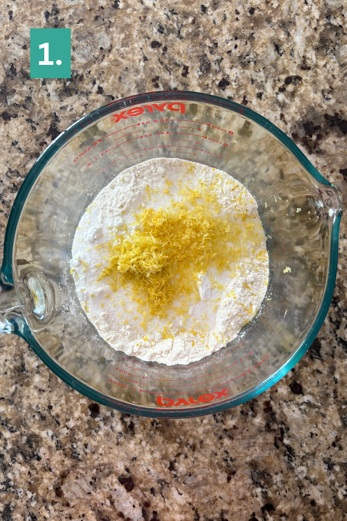 A glass measuring cup with flour and lemon zest, ready for lemon blueberry scones, sits on a granite countertop, labeled with a green “1” in the top left corner.