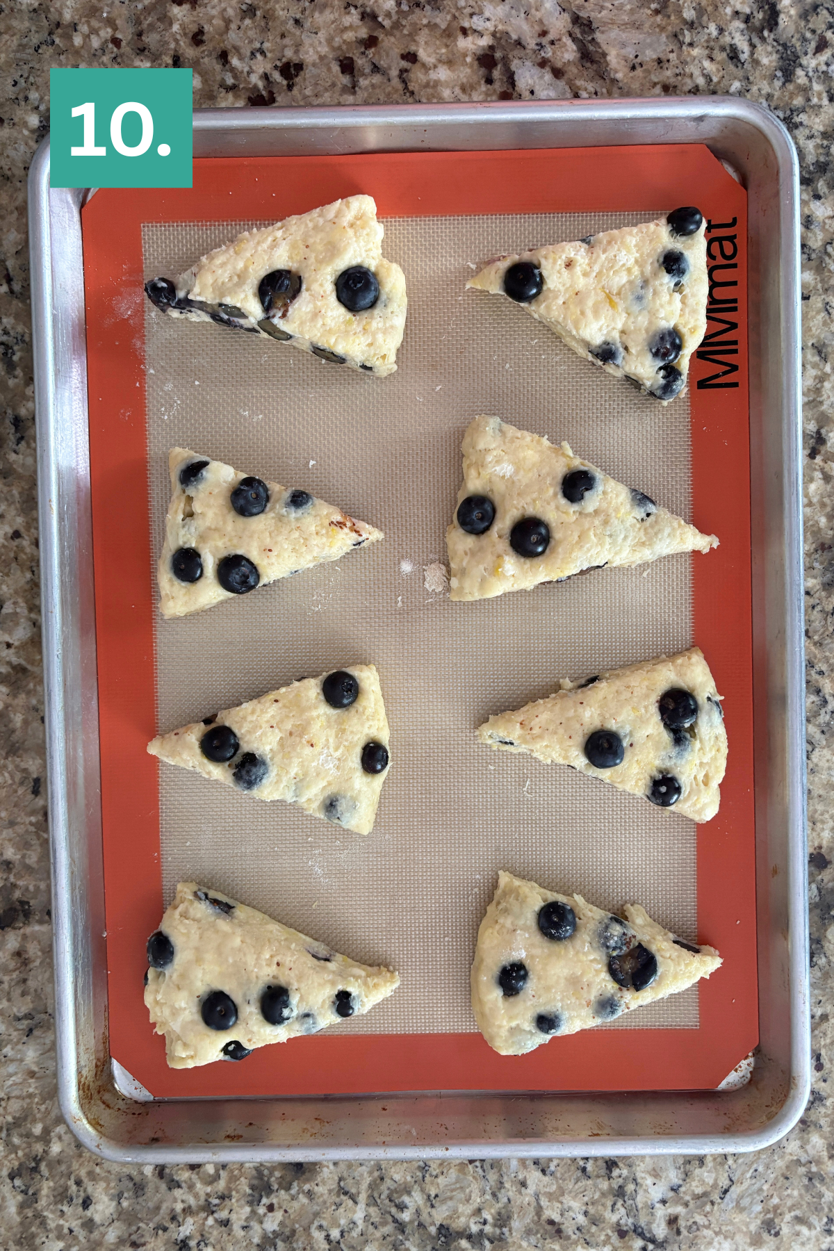 A baking sheet with eight unbaked, triangular lemon blueberry scones on a silicone mat rests on a granite countertop. A green label with the number “10.” appears in the top left corner.