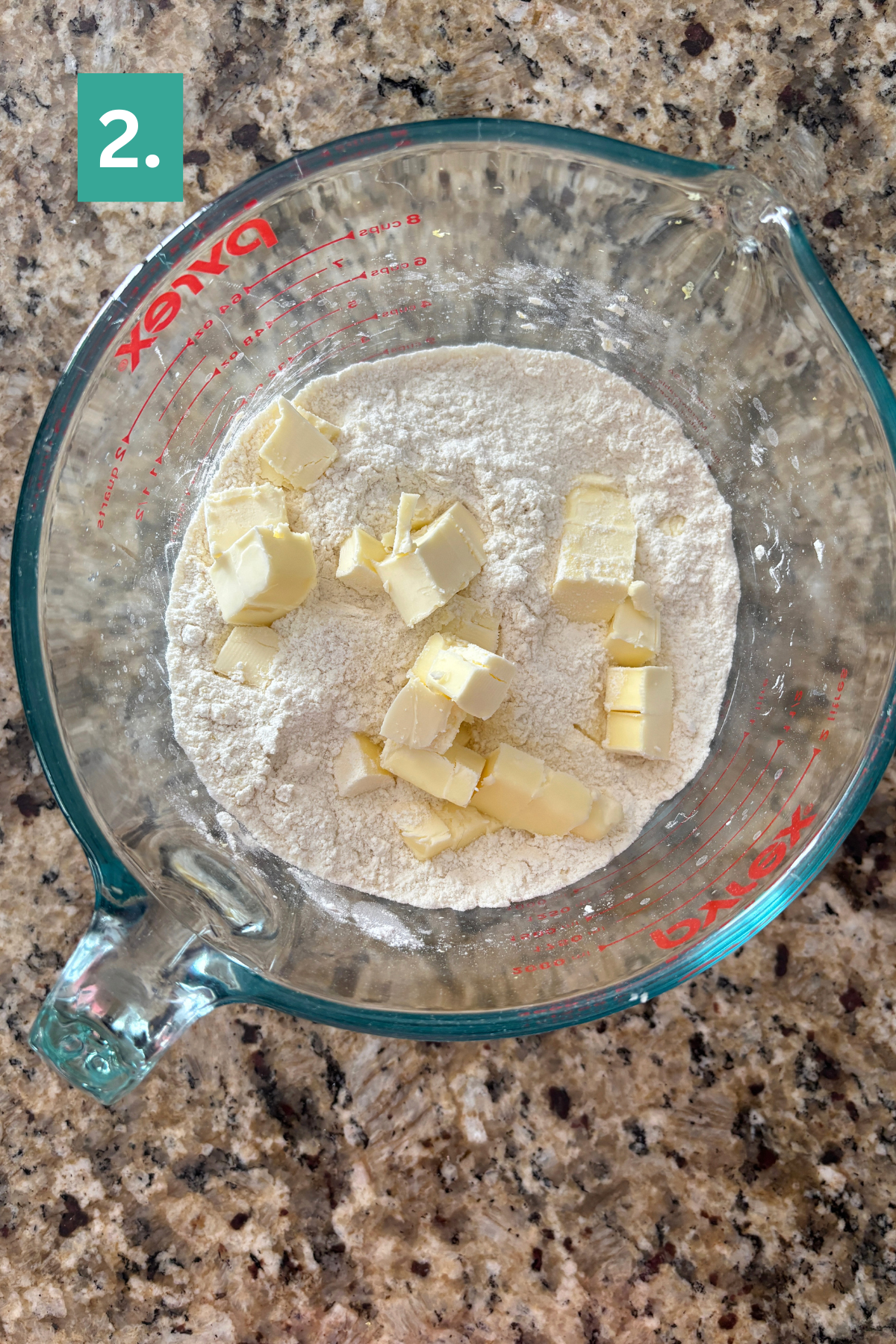 A glass measuring cup filled with flour and cubes of butter sits on a granite countertop—ingredients for lemon blueberry scones. A green square with the number 2. appears in the top left corner.