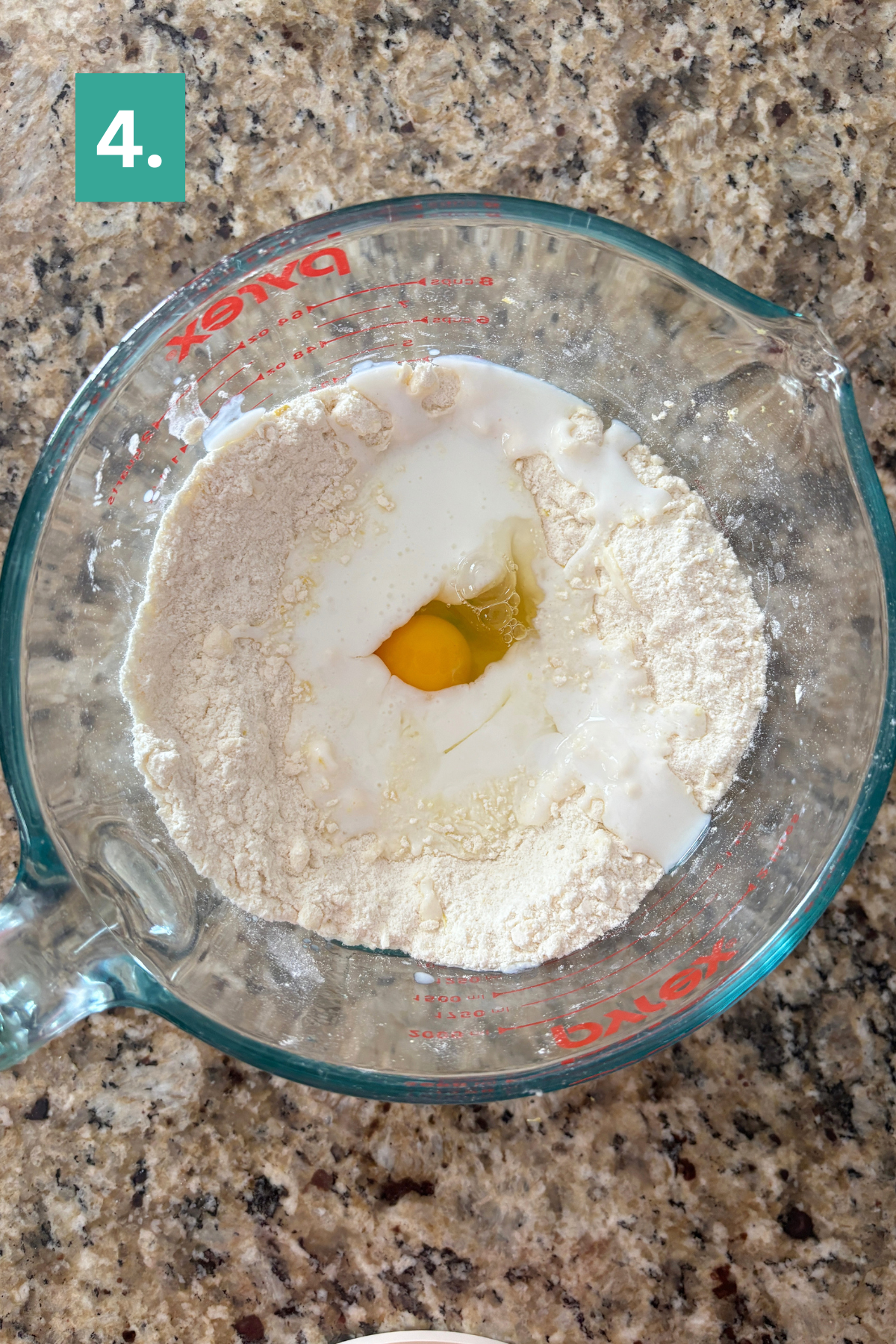 A glass measuring cup containing flour, milk, and a raw egg sits on a granite countertop—perfect ingredients for lemon blueberry scones. A green box with the number 4 appears in the top left corner of the image.