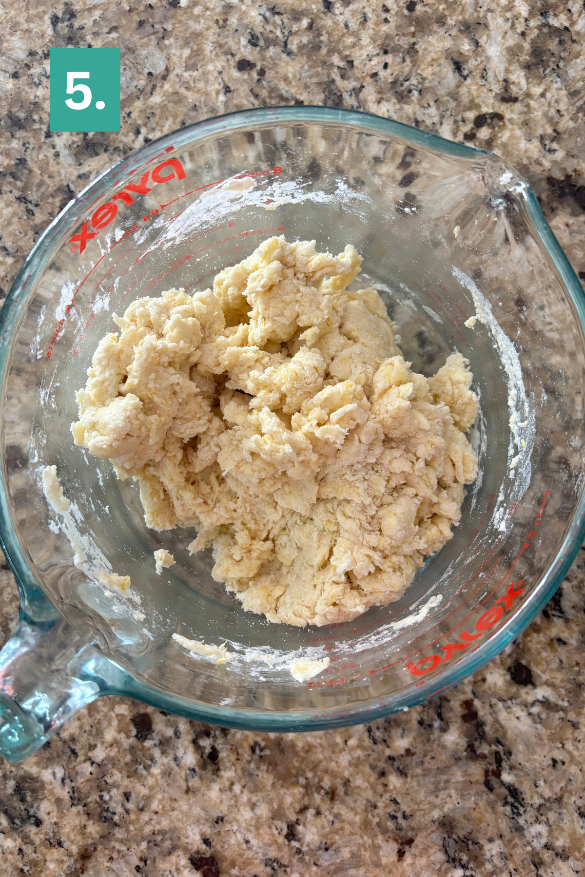 A glass measuring cup filled with a rough, slightly crumbly ball of lemon blueberry scone dough sits on a granite countertop. A green box with the number 5 appears in the top left corner of the image.