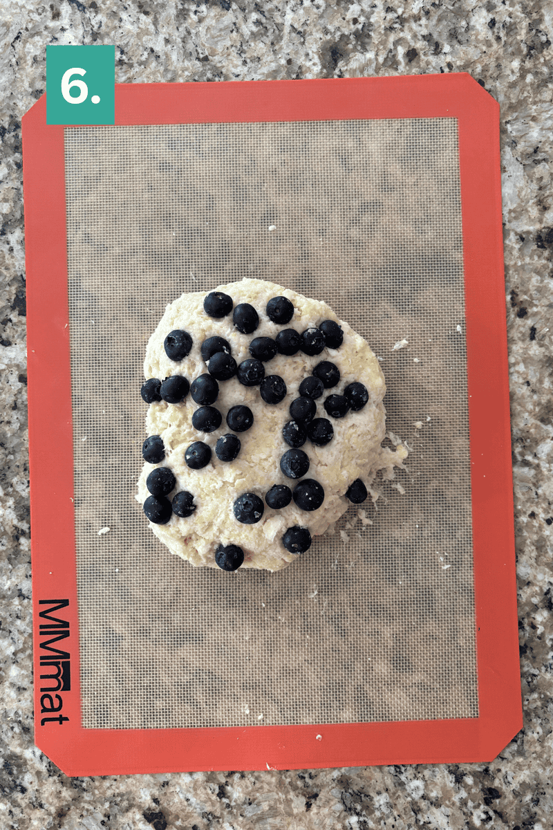 A mound of dough for lemon blueberry scones, topped with fresh blueberries, sits on a silicone baking mat on a granite countertop. A green box with the number 6 appears in the top left corner.