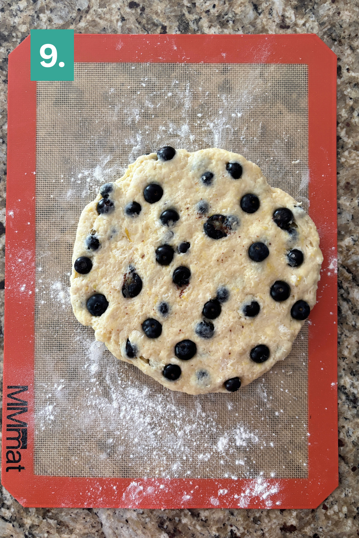 Round scone dough for lemon blueberry scones with fresh blueberries rests on a lightly floured silicone baking mat atop a granite countertop. A green box with the number 9 appears in the top left corner.