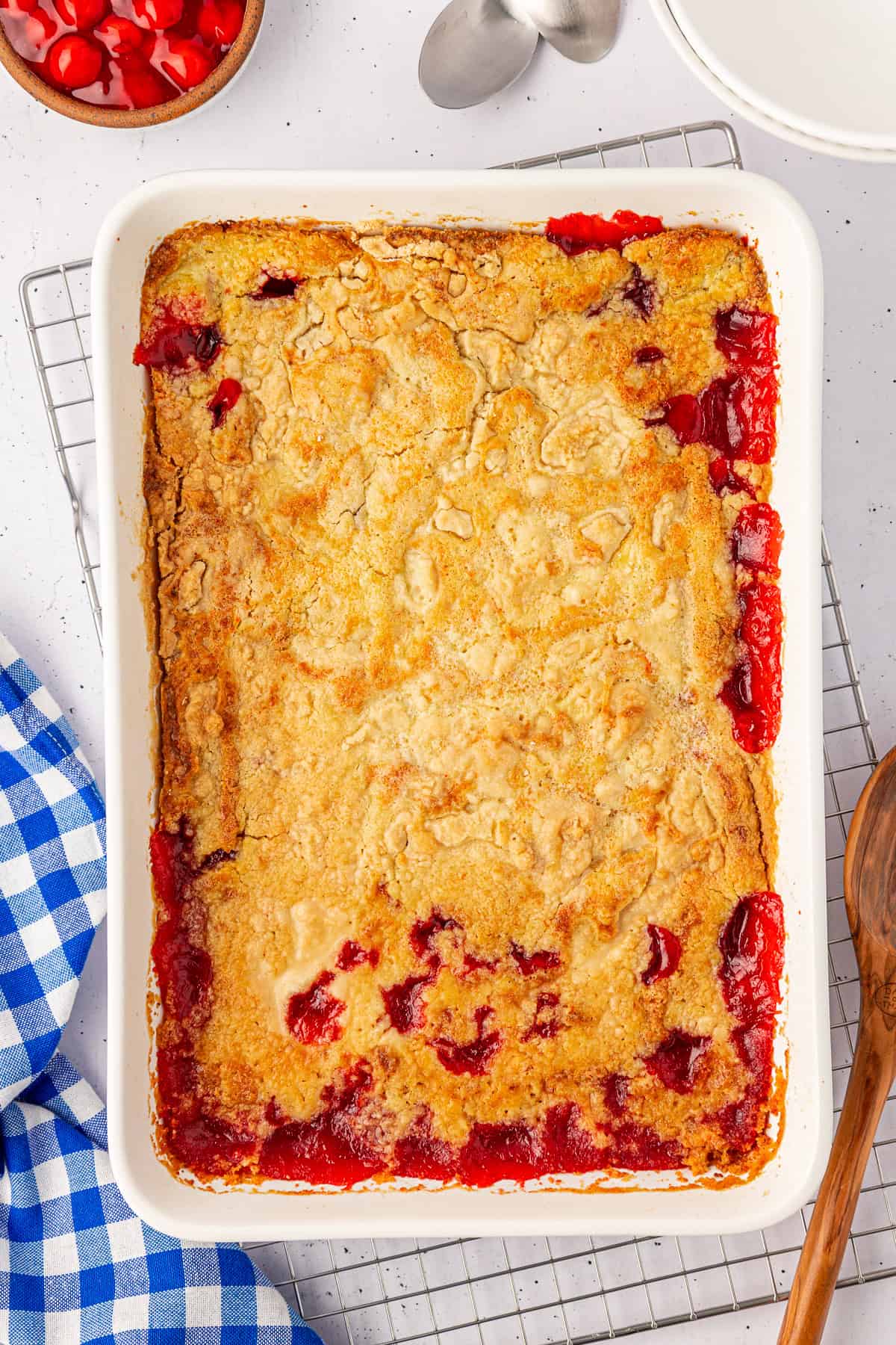 A rectangular baking dish holds a delectable cherry cobbler with a golden crust and red cherry filling peeking through the edges. Alongside, there’s a blue and white checkered cloth, a wooden spoon, and a bowl of cherries—reminiscent of the classic cherry dump cake.