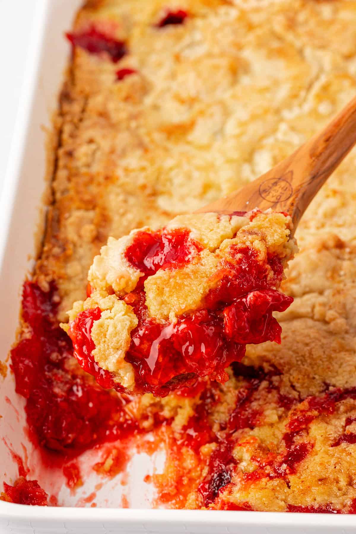 A close-up of a wooden spoon scooping a serving of cherry dump cake from a white baking dish. The dish showcases a golden-brown crust with vibrant red cherry filling bubbling below, giving it a warm and inviting appearance.