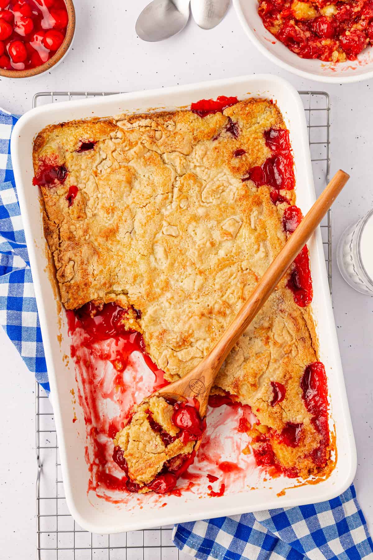 A white baking dish filled with cherry dump cake, partially eaten, rests on a cooling rack. A wooden spoon is placed on top. Two empty silver spoons and a small checkered blue cloth lie nearby. In the background, a glass of milk and a small bowl of cobbler complete the inviting scene.