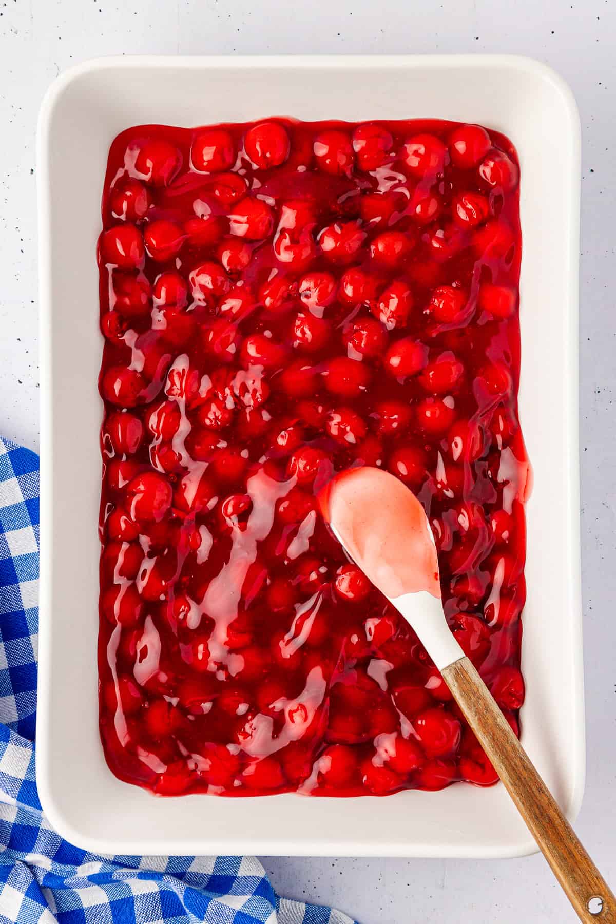 A white baking dish brims with glistening red cherry pie filling, hinting at a delicious cherry dump cake. A white spatula with a wooden handle rests on the edge of the dish, while a blue and white checkered cloth peeks out from the bottom left corner.